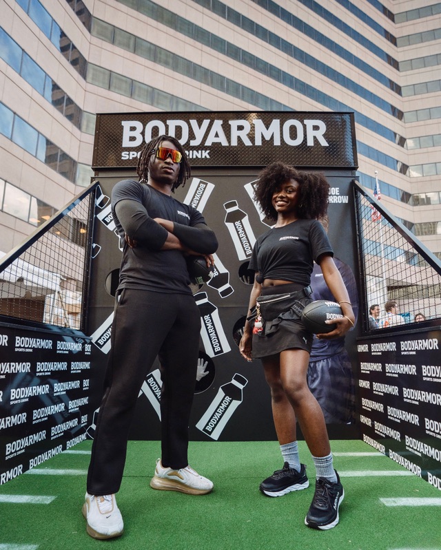 Two BodyArmor brand ambassadors at the branded football-toss game wall on an elevated turf lane at the Kroger Wellness Festival