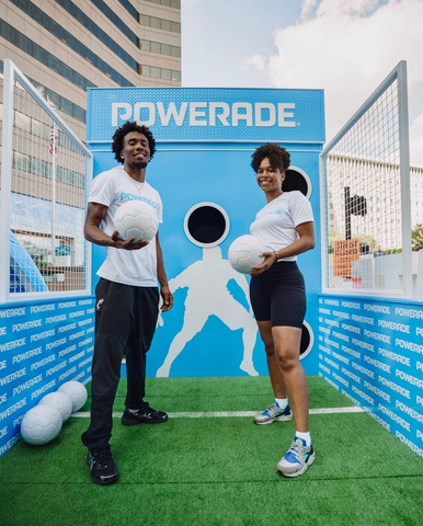 Two Powerade brand ambassadors holding soccer balls at the branded soccer-toss game wall on the turf lane at the Kroger Wellness Festival